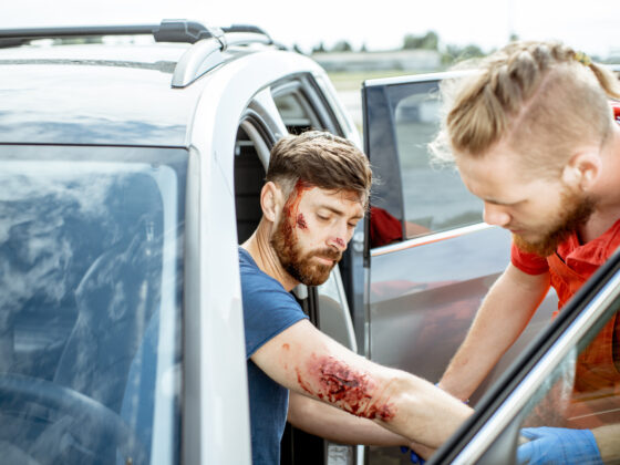 Ambulance worker examining injuries of a man sitting near the car after the road accident, providing emergency medical assistance